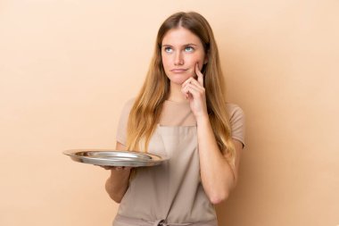 Tepsisi bej arka planda olan genç sarışın garson kadın yukarı bakarken şüpheleri var.