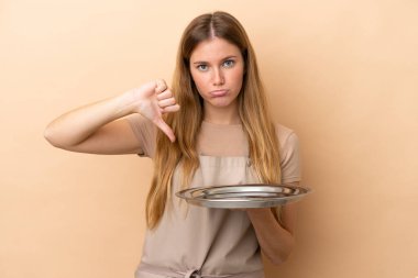 Bej arka planda izole edilmiş tepsisi olan genç sarışın garson kadın olumsuz bir ifadeyle baş parmağını aşağı gösteriyor.