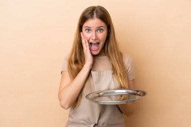 Tepsisi bej arka planda, şaşırmış ve şok olmuş bir yüz ifadesi olan genç sarışın garson kadın.