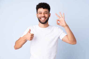 Faslı genç bir adam mavi arka planda izole edilmiş. Tamam işareti gösteriyor.