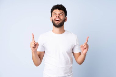 Faslı genç adam mavi arka planda izole edilmiş, şaşırmış ve yukarı bakıyor.