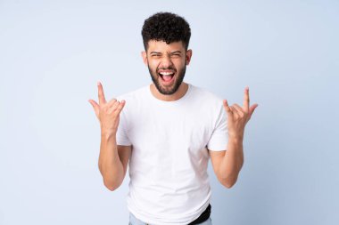 Faslı genç bir adam mavi arka planda izole edilmiş boynuz hareketi yapıyor.