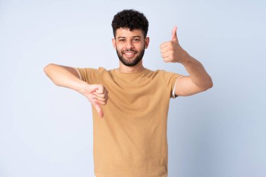 Faslı genç bir adam mavi arka planda izole edilmiş, iyiye kötüye işaret yapıyor. Evet ya da hayır arasında kararsız