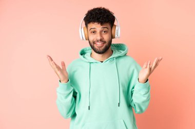 Faslı genç bir adam pembe arka planda izole edilmiş şaşırmış ve müzik dinliyor.