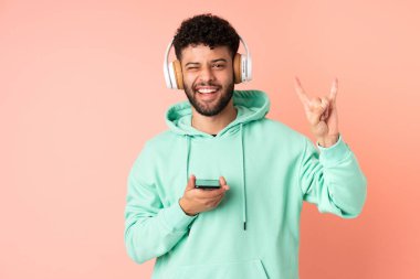 Genç Faslı bir adam pembe arka planda izole edilmiş, hareketli bir rock hareketi yaparak müzik dinliyor.
