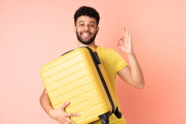 Genç Faslı bir adam pembe arka planda izole edilmiş seyahat valiziyle geziyor ve 