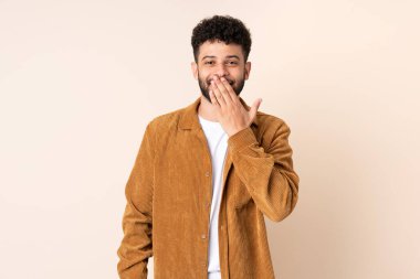 Faslı genç bir adam bej arka planda izole edilmiş mutlu ve gülümseyen ağızlarını eliyle kapatıyor.