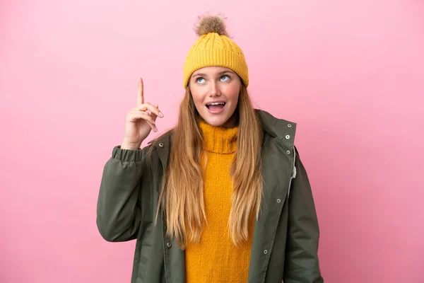 年轻的金发女人穿着冬季夹克 背景是粉色的 想在举起一只手指头的同时实现这个解决方法 — 图库照片