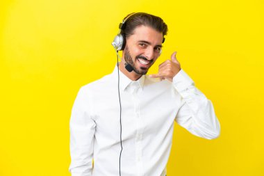 Tele pazarlamacı beyaz adam sarı arka planda izole edilmiş bir kulaklıkla çalışıyor ve telefon jesti yapıyor. Bana geri dönüş işareti ver.