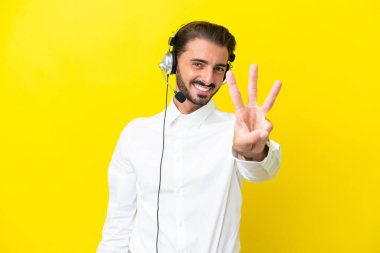 Tele pazarlamacı beyaz adam sarı arka planda izole edilmiş bir kulaklıkla çalışıyor mutlu ve parmaklarıyla üçe kadar sayıyor.