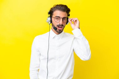 Tele pazarlamacı beyaz adam sarı arka planda gözlüklü ve şaşırmış bir kulaklıkla çalışıyor.
