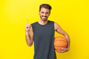 Yakışıklı genç basketbolcu sarı arka planda izole edilmiş ve en iyinin işareti olarak parmağını kaldırıyor.
