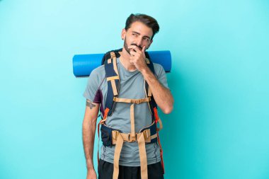 Mavi arka planda izole edilmiş büyük bir sırt çantası olan dağcı genç adamın şüpheleri ve kafa karıştırıcı bir yüz ifadesi var.