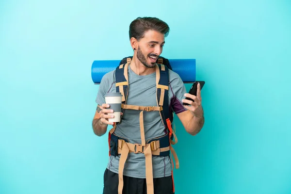 若い登山家の男と大きなバックパックの青の背景に隔離されたコーヒーを取るとモバイルを保持 — ストック写真