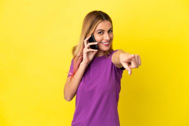 İzole edilmiş sarı arka plan üzerinde cep telefonu kullanan genç bir kadın mutlu bir ifadeyle ön tarafı işaret ediyor.