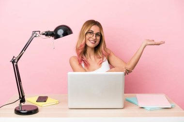 İşyerindeki pembe arka planda dizüstü bilgisayarı olan genç bir bayan davet edildiği için ellerini yana uzatıyor.