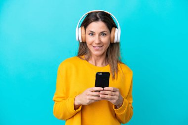 Orta yaşlı beyaz bir kadın mavi arka planda izole edilmiş, cep telefonuyla müzik dinliyor ve ön tarafa bakıyor.