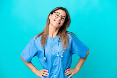 Mavi arka planda izole edilmiş orta yaşlı cerrah kadın kolları kalçasında ve gülümsüyor.