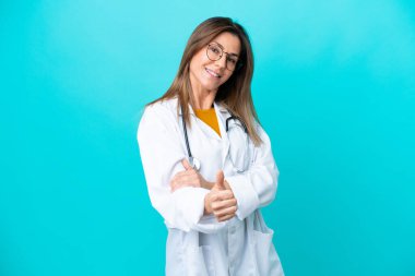 Mavi arka planda izole edilmiş orta yaşlı bir kadın doktor önlüğü giyiyor ve başparmağı yukarıda.