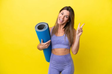 Spor kadını yoga derslerine giderken elinde sarı olmayan bir minder tutarken gülümsüyor ve zafer işareti gösteriyor.