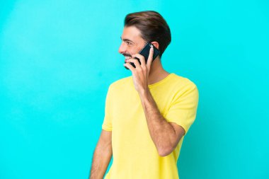 Kafkasyalı genç adam mavi arka planda izole edildi. Cep telefonuyla biriyle sohbet ediyor.