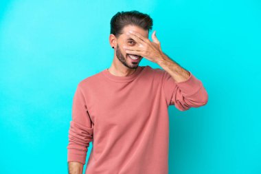 Mavi arka planda izole edilmiş genç beyaz bir adam elleriyle gözlerini kapatıyor ve gülümsüyor.