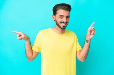 Mavi arka planda izole edilmiş genç beyaz adam parmak uçlarını işaret ediyor ve mutlu.