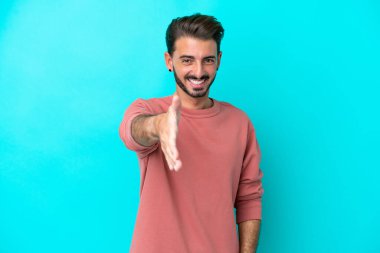 Mavi arka planda izole edilmiş genç beyaz adam iyi bir anlaşma için el sıkışıyor.