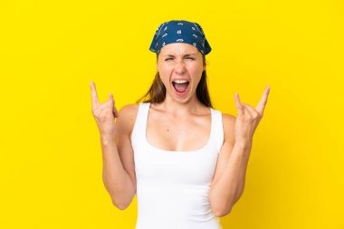 Sarı arka planda izole edilmiş genç bir İngiliz kadın boynuz hareketi yapıyor.