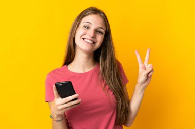 Sarı arka planda izole edilmiş cep telefonu kullanan genç kadın gülümsüyor ve zafer işareti gösteriyor.