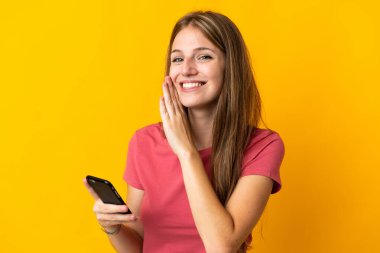 Sarı arka planda izole edilmiş cep telefonu kullanan genç kadın bir şeyler fısıldıyor.