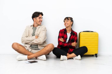 Genç melez ırk gezginleri, beyaz arka planda izole bir şekilde yerde oturuyor. Gülümseyerek omuzlarının üzerinden bakıyorlar.