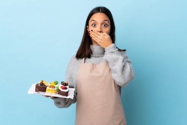 Pasta şefi, izole edilmiş mavi arka plan üzerinde büyük bir pasta tutuyor. Ağzını ellerle kapatıyor..