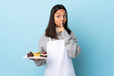 Pasta şefi, izole edilmiş mavi arka planda büyük bir pasta tutuyor. Parmağını ağzına koyduğunda sessizlik işareti gösteriyor..