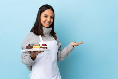Pasta şefi mavi arka planda büyük bir pasta tutuyor. Bir yandan gülümserken bir fikir sunuyor..