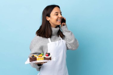 Pasta şefi, mavi arka planda büyük bir pasta tutuyor cep telefonuyla sohbet ediyor..