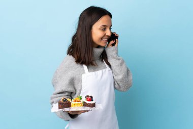 Pasta şefi, mavi arka planda büyük bir pasta tutuyor ve cep telefonuyla sohbet ediyor..