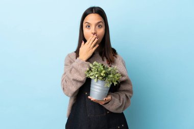 Genç, esmer, melez bir kadın mavi arka planda bir bitki tutuyor sağa bakarken şaşırmış ve şok olmuş..