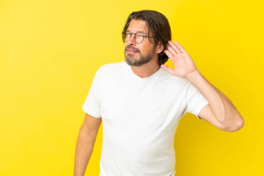 Yaşlı Hollandalı adam sarı arka planda izole edilmiş. Kulağına elini sürerek bir şeyler dinliyor.