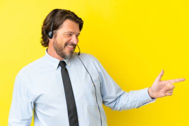 Tele pazarlamacı Hollandalı adam sarı arka planda izole edilmiş bir kulaklıkla çalışıyor. Parmakla işaret ediyor ve bir ürün sunuyor.