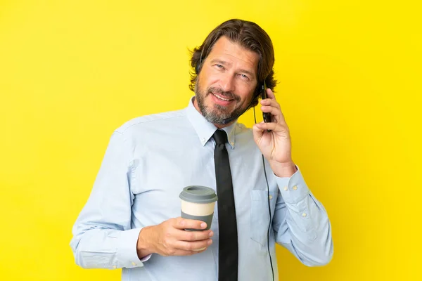 电话推销员带着耳机工作 耳机孤立在黄色的背景下 手里拿着咖啡 一边拿着手机 — 图库照片