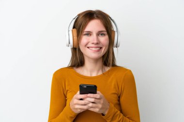 Genç bir İngiliz kadın beyaz arka planda izole edilmiş. Cep telefonuyla müzik dinliyor ve ön tarafa bakıyor.