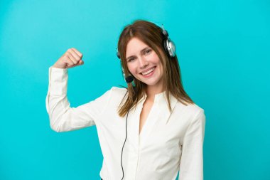 Tele pazarlamacı İngiliz kadın mavi arka planda izole edilmiş bir kulaklıkla çalışıyor. Güçlü bir jest yapıyor.