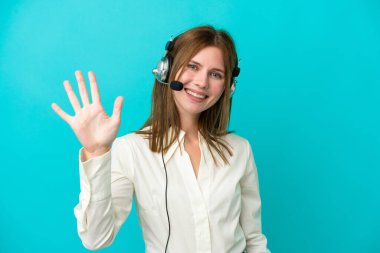 Tele pazarlamacı İngiliz kadın mavi arka planda izole edilmiş kulaklıkla çalışıyor parmaklarıyla beşi sayıyor.