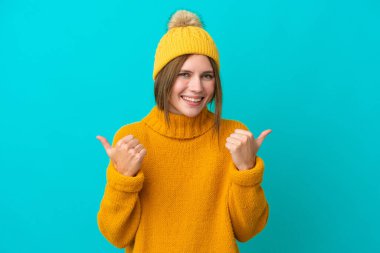 Kışlık ceket giyen genç İngiliz kadın mavi arka planda izole edilmiş başparmakları havada ve gülümsüyor.