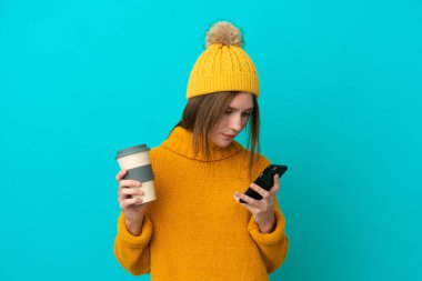 Kışlık ceket giyen genç İngiliz kadın mavi arka planda izole bir şekilde kahve ve bir cep telefonu tutuyor.