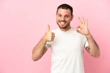 Pembe arka planda izole edilmiş genç Brezilyalı adam işaret ve baş parmak hareketi gösteriyor.