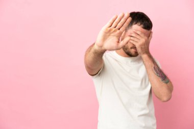 Pembe arka planda izole edilmiş genç Brezilyalı adam jesti durdurup yüzünü kapatıyor.