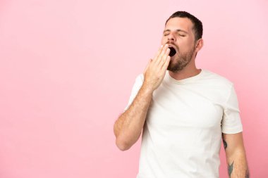 Pembe arka planda izole edilmiş genç Brezilyalı bir adam esniyor ve ağzını eliyle açık tutuyor.