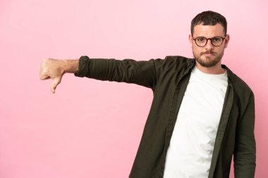 Pembe arka planda izole edilmiş genç Brezilyalı adam olumsuz ifadeyle baş parmağını aşağı gösteriyor.
